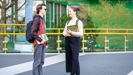 [Translate to Englisch:] Studierende unterhalten sich auf dem Campus mit Büchern in der Hand. 